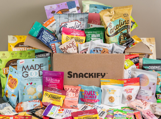 Assorted snack packets in front of a Snackify box on a neutral background